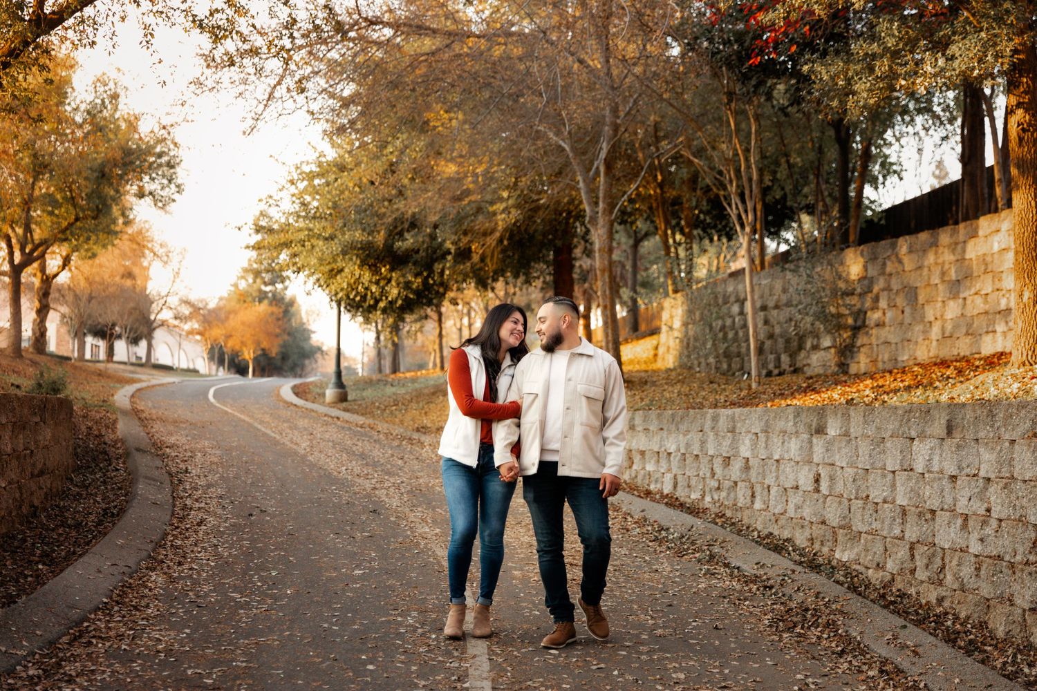 Couple walking together on a tree-lined autumn path with golden leaves and stone wall.