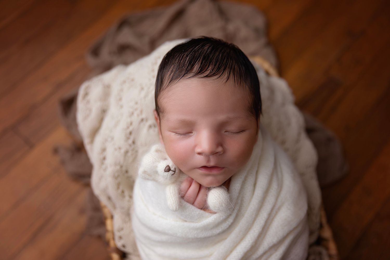 Peaceful newborn wrapped in white with small teddy bear on wooden floor.