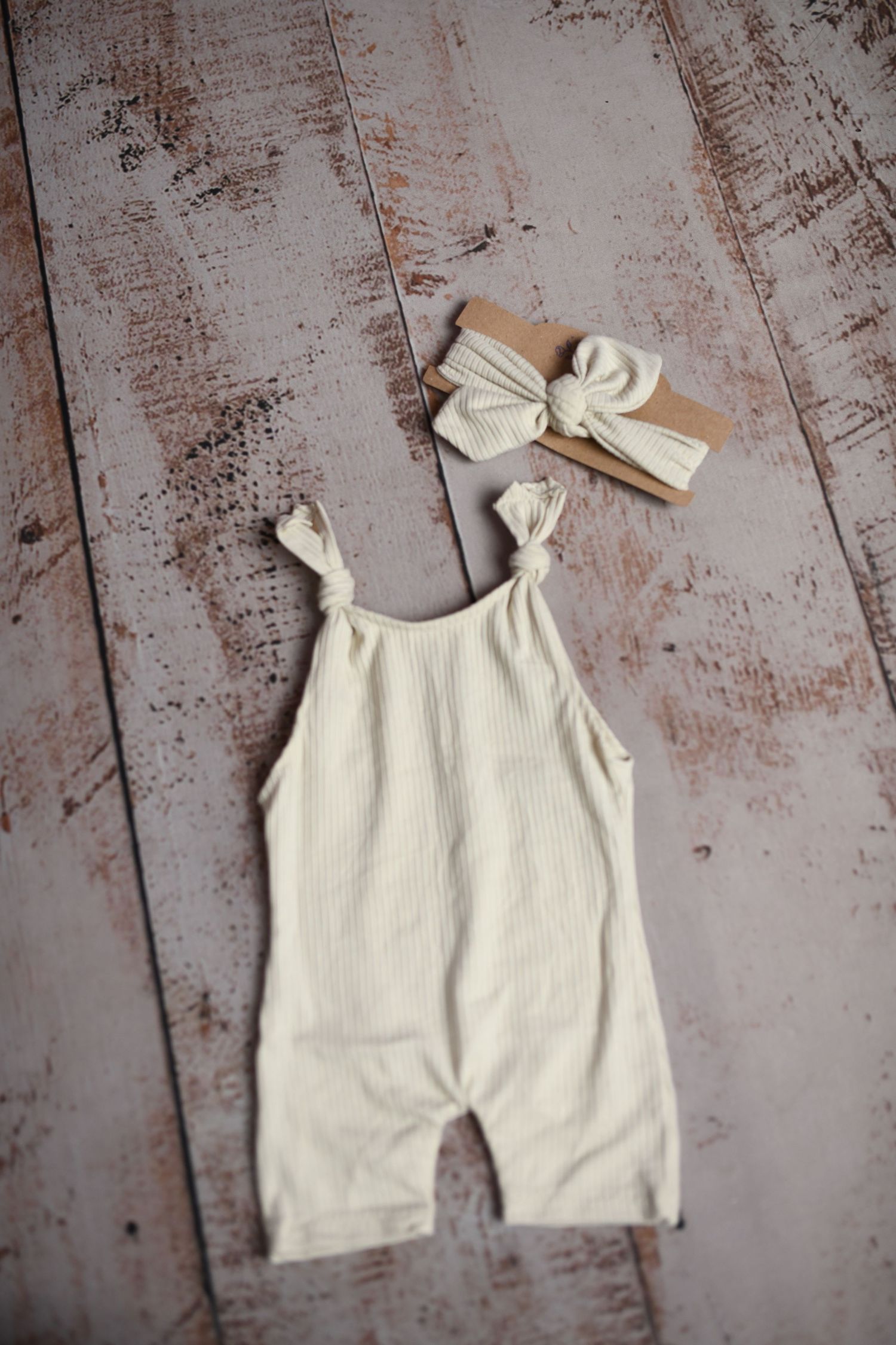 Cream colored ribbed romper with bow ties displayed on distressed wooden background.