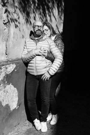 Black and white portrait of couple in matching puffer jackets leaning against textured wall.