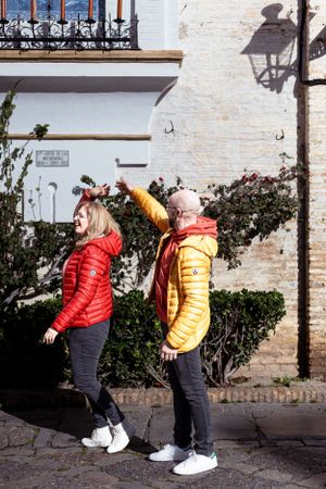 Two people in bright puffy jackets posing playfully against a white wall with ornate balcony.