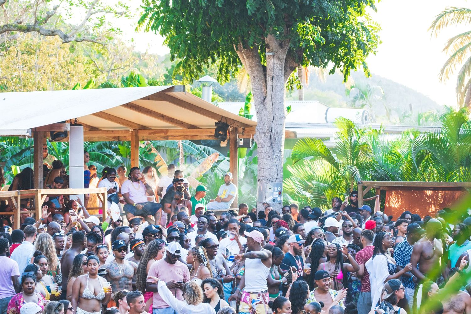 Large crowd gathering under a wooden canopy structure surrounded by tropical palm trees and greenery at an outdoor event.