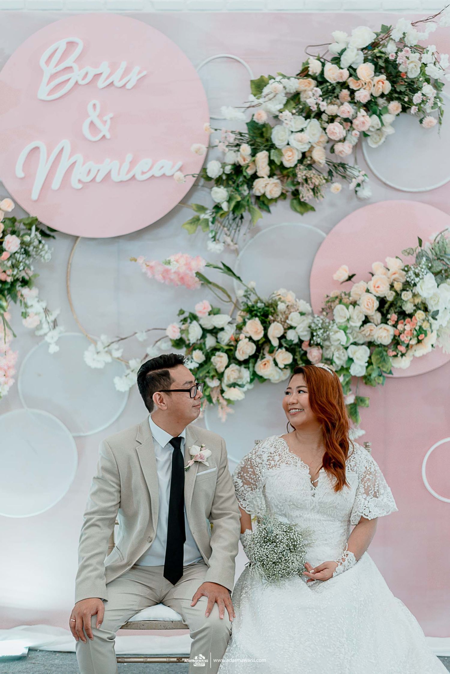 Couple sharing intimate moment on decorated bench against pink circular backdrop with floral details.