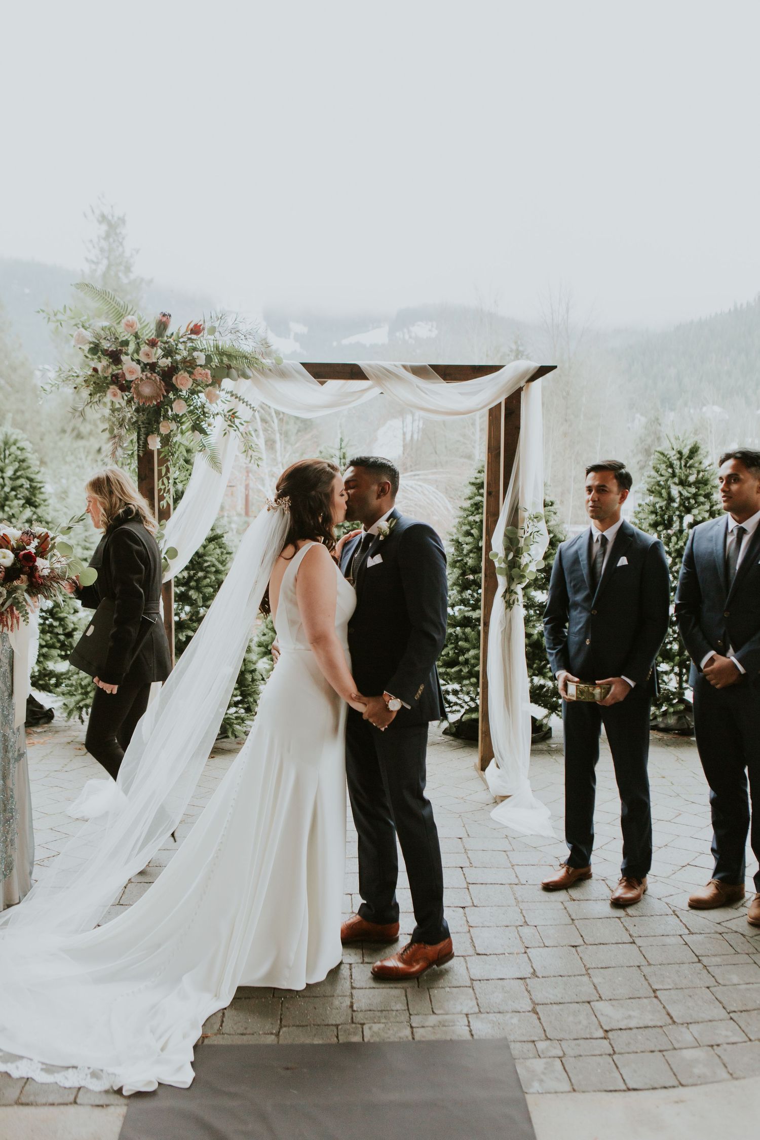 A romantic outdoor wedding ceremony takes place under a white draped pergola with mountain views and floral arrangements.