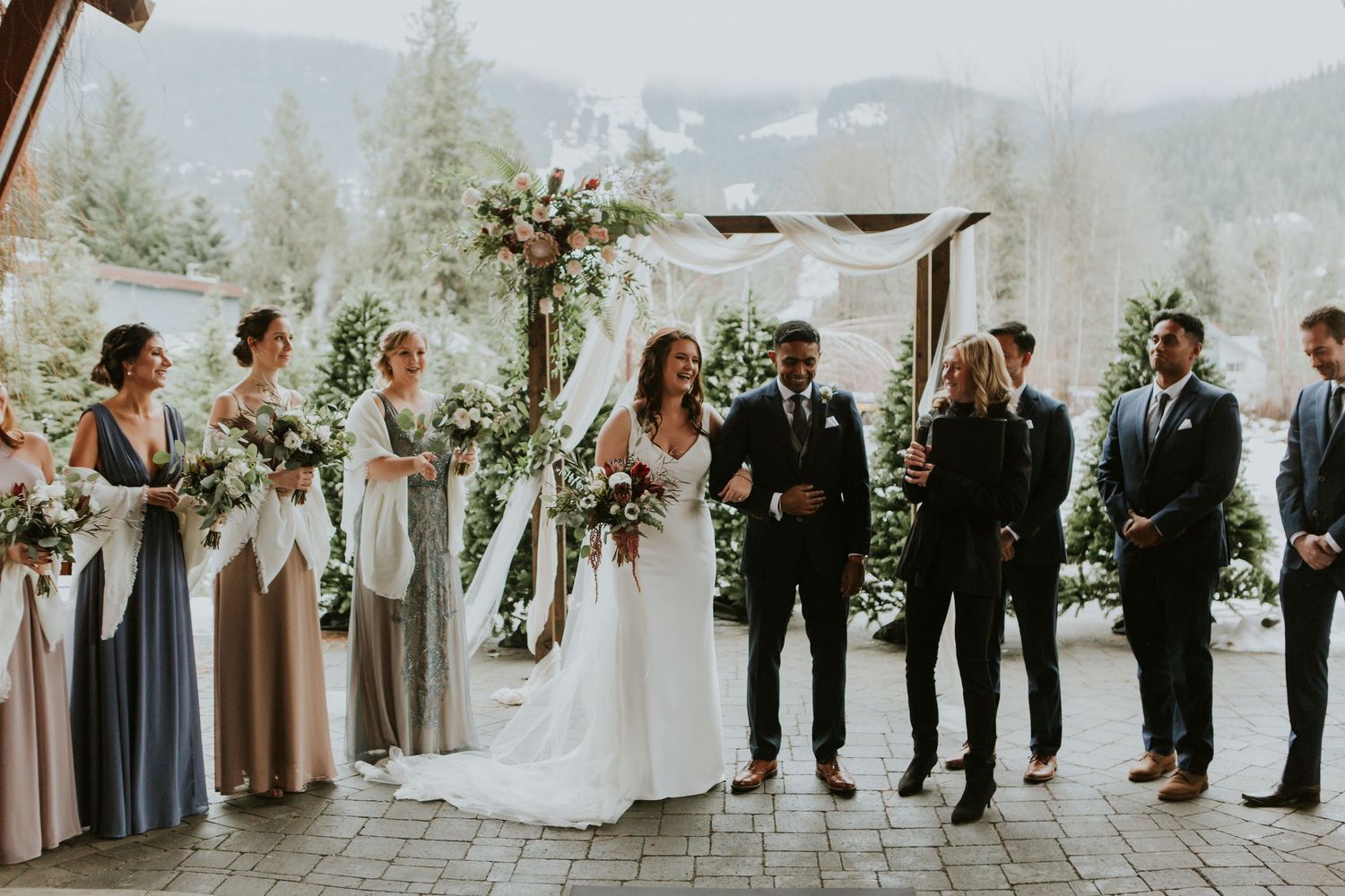 Elegant outdoor mountain wedding ceremony under a white canopy with bridal party in blue and blush tones.