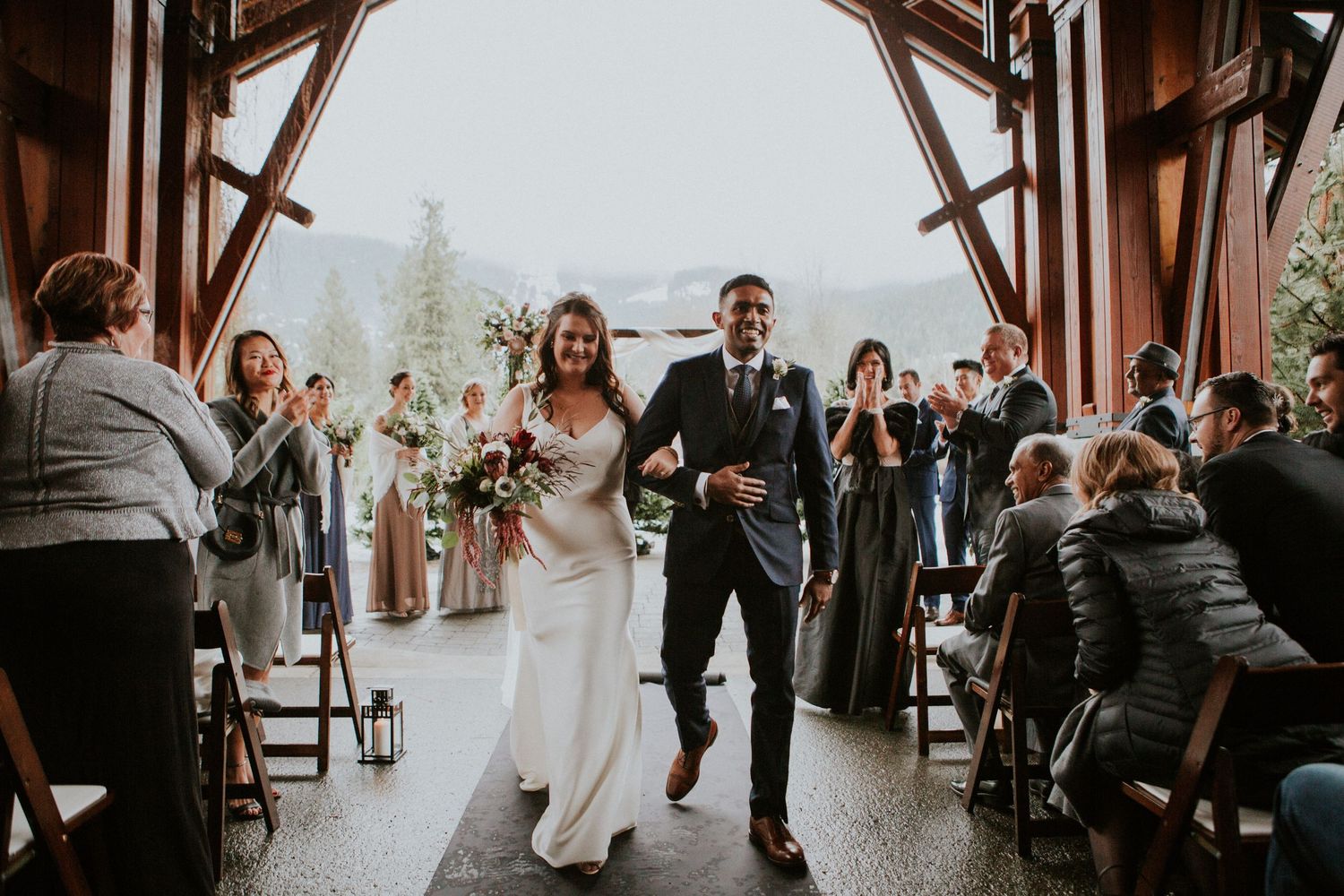 Happy couple exits rustic wooden wedding ceremony venue surrounded by cheering guests and holding hands.