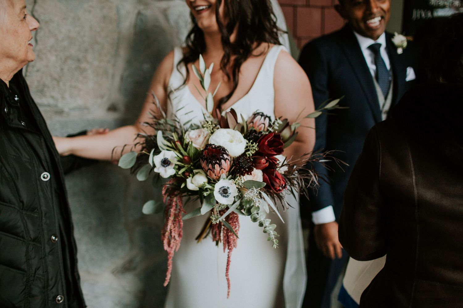 A wedding bouquet with white anemones, burgundy flowers and trailing greenery is held against a white dress.