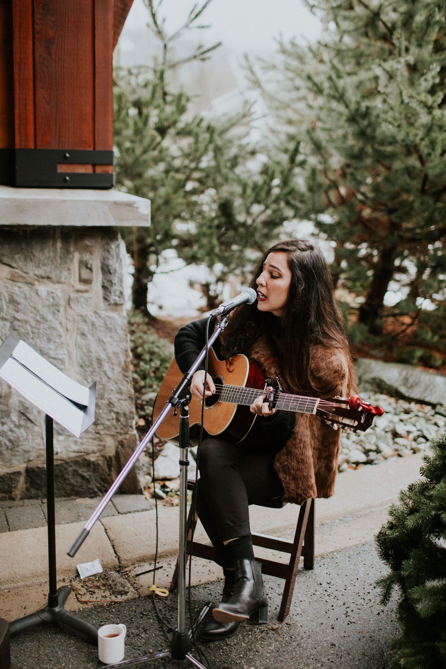 Musician performs acoustic guitar and sings into microphone while seated outdoors by stone wall and evergreen trees.
