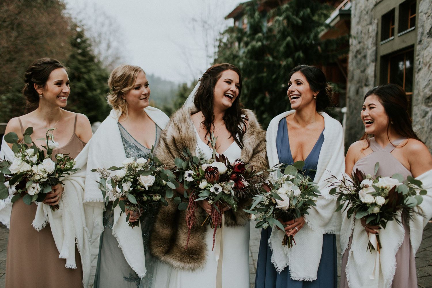 A group of wedding party members share a candid laughing moment while holding beautiful floral bouquets.