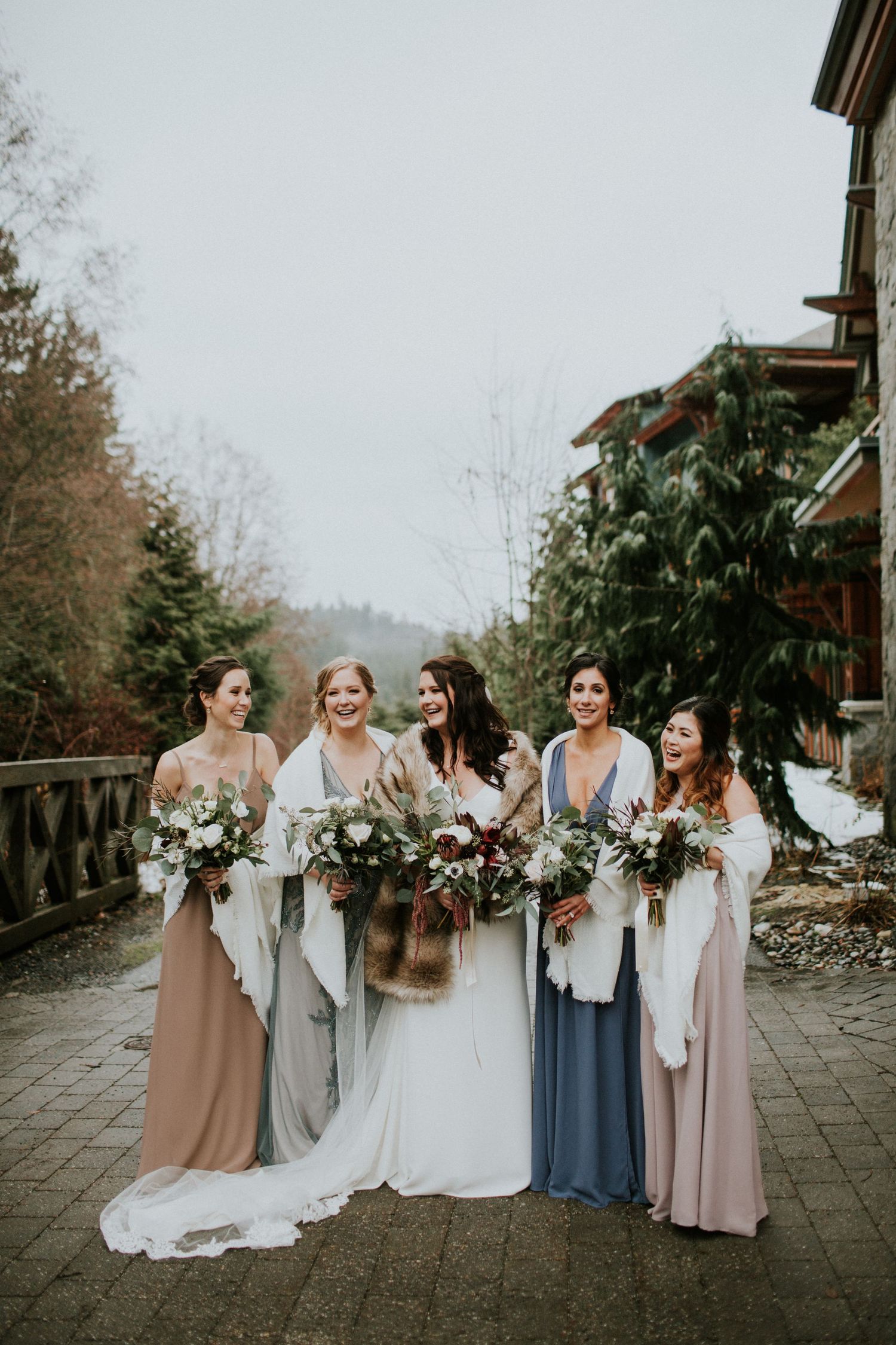 Winter wedding party in soft pastel dresses holds bouquets while standing on stone patio at mountain resort.