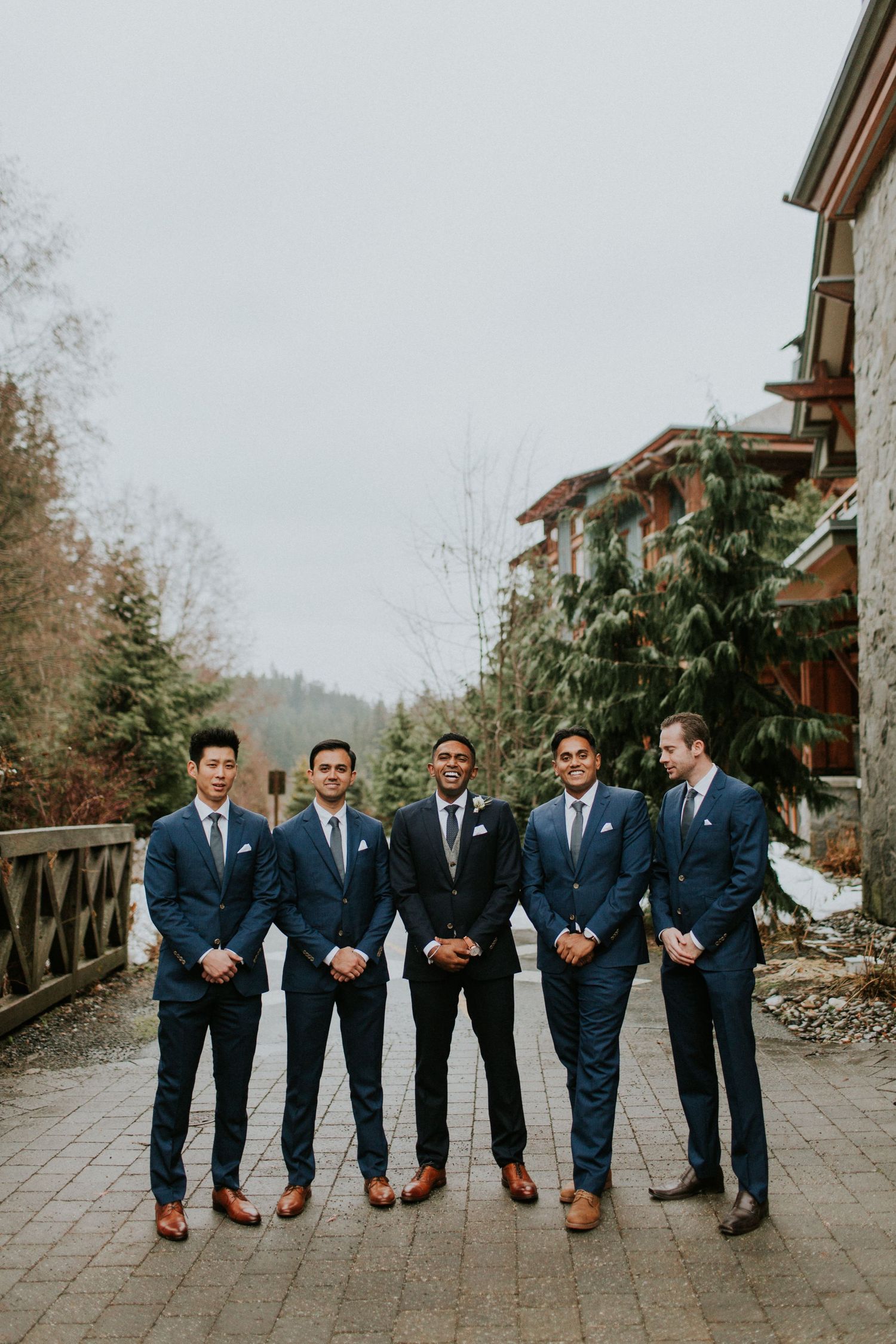 A group of groomsmen wearing coordinated navy suits and brown shoes pose together on a paved path at a mountain resort.