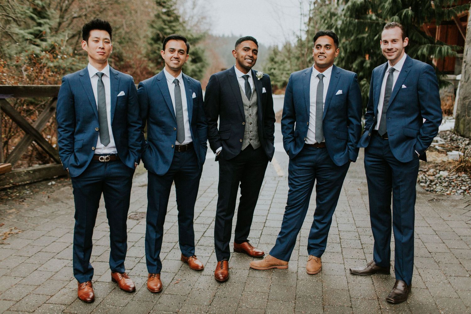Five groomsmen wearing matching navy suits with light blue ties stand together on a paved outdoor path.