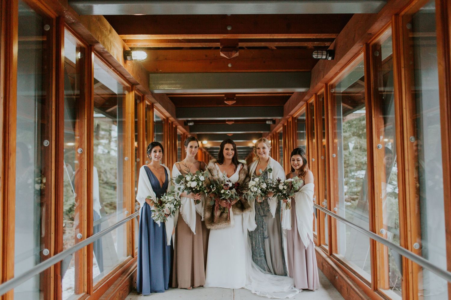 A wedding party stands together in a rustic wooden covered walkway holding neutral floral bouquets.