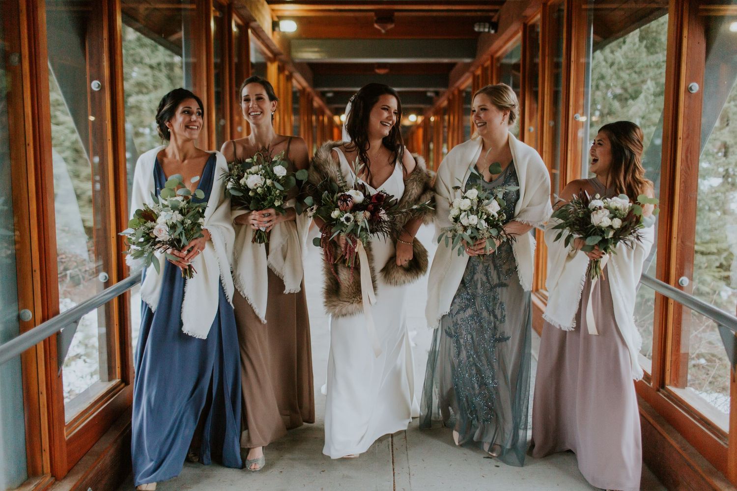 Bridal party in mismatched pastel dresses holding white floral bouquets pose together in rustic venue hallway.