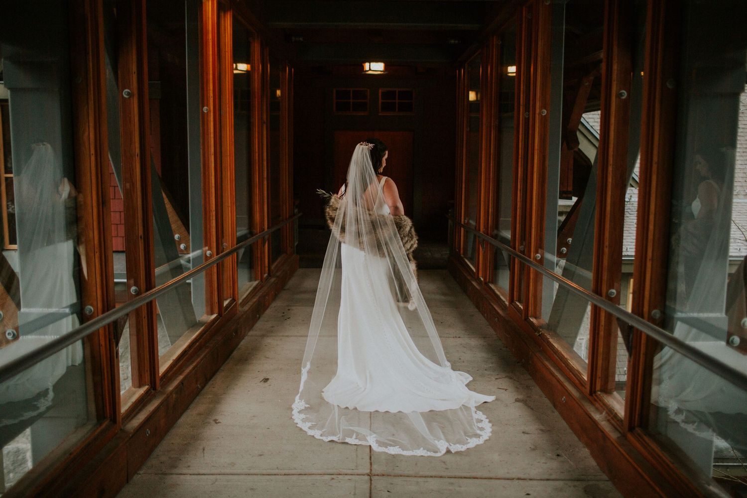 A bride in a flowing white gown and veil stands in a long hallway with warm wooden beams and glass panels.