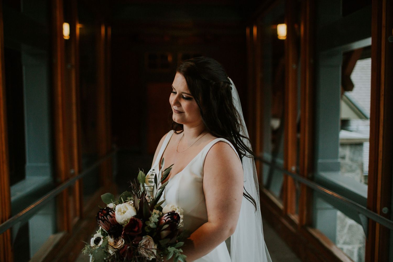 Bride in white dress holding dark floral bouquet stands in dimly lit hallway with wooden windows.