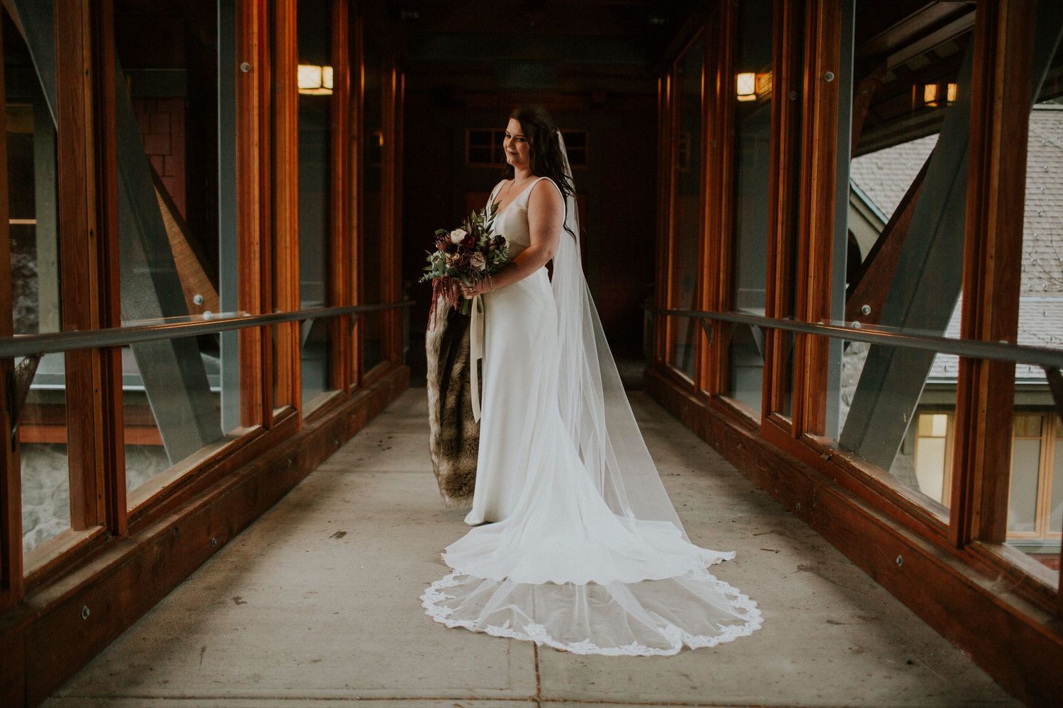 A bride in a flowing white gown and long veil stands in a warmly lit wooden hallway with glass windows.