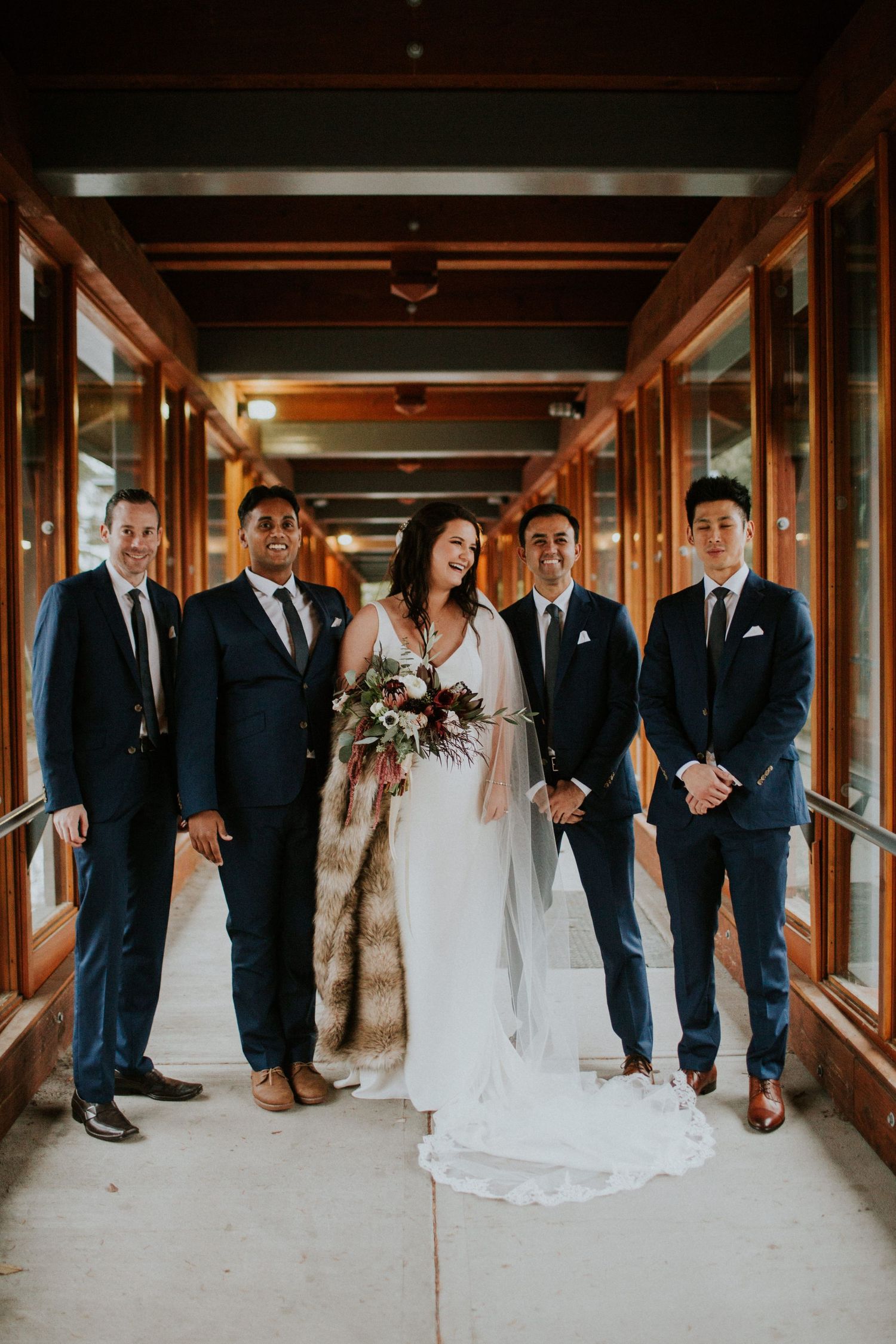 Wedding party poses together in elegant formal attire inside a wood-paneled hallway with natural lighting.