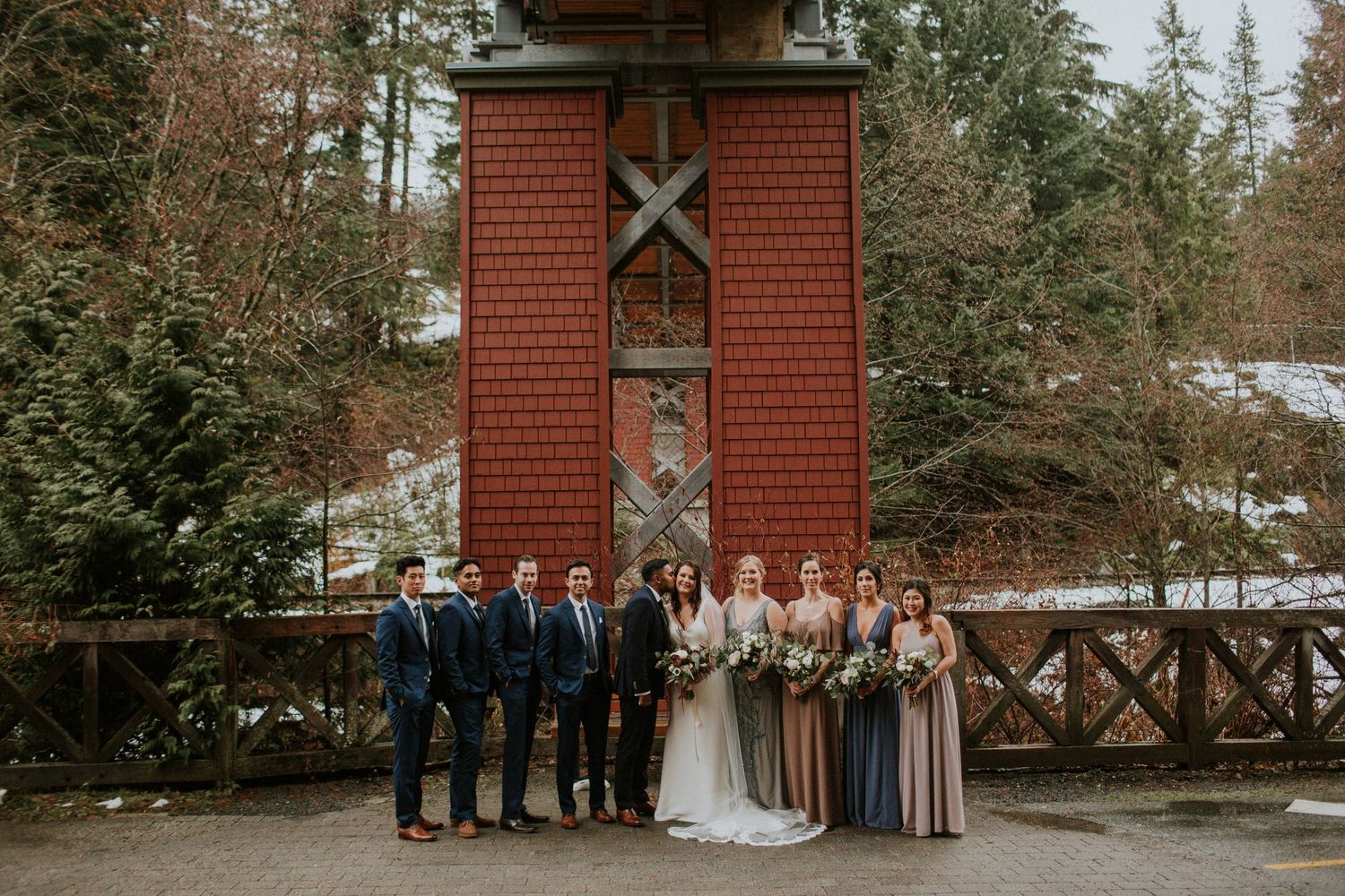 Wedding party poses in front of rustic red wooden tower structure surrounded by evergreen trees in winter setting.