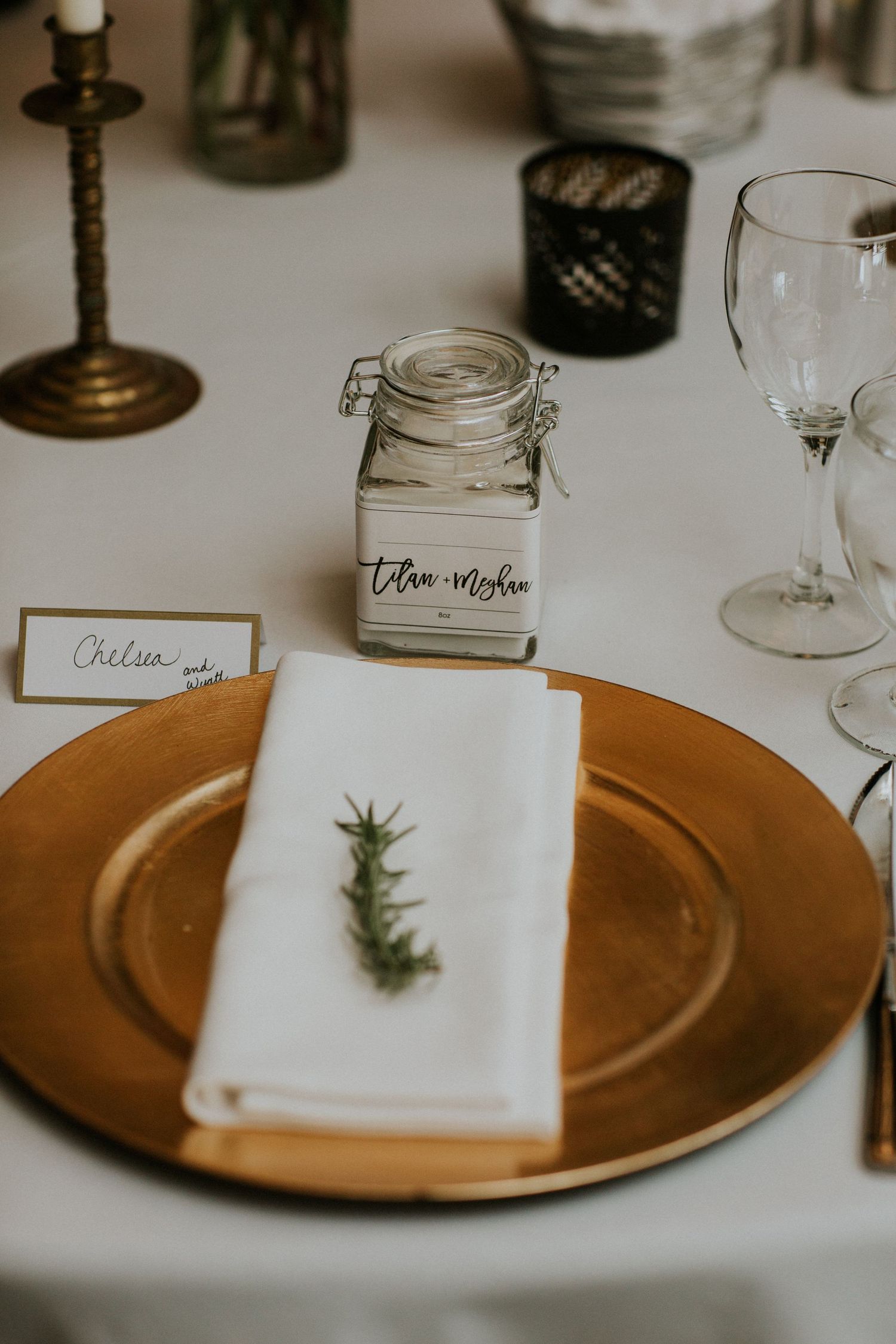 Elegant wedding place setting with gold charger plate, white napkin, rosemary sprig, and glass jar table decor.