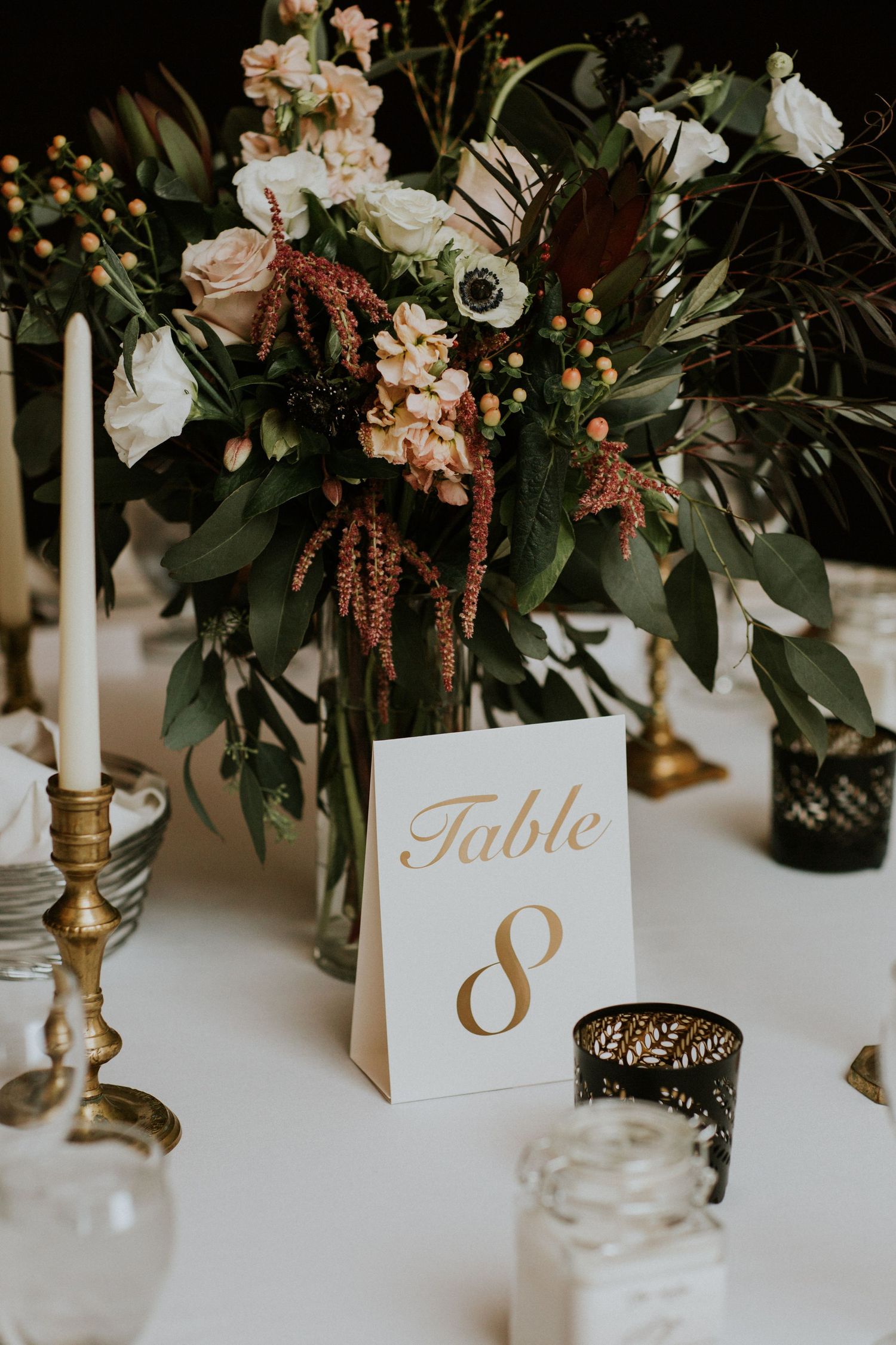 Elegant wedding reception table decor with floral centerpiece, gold table number 8, and white taper candles in brass holders.