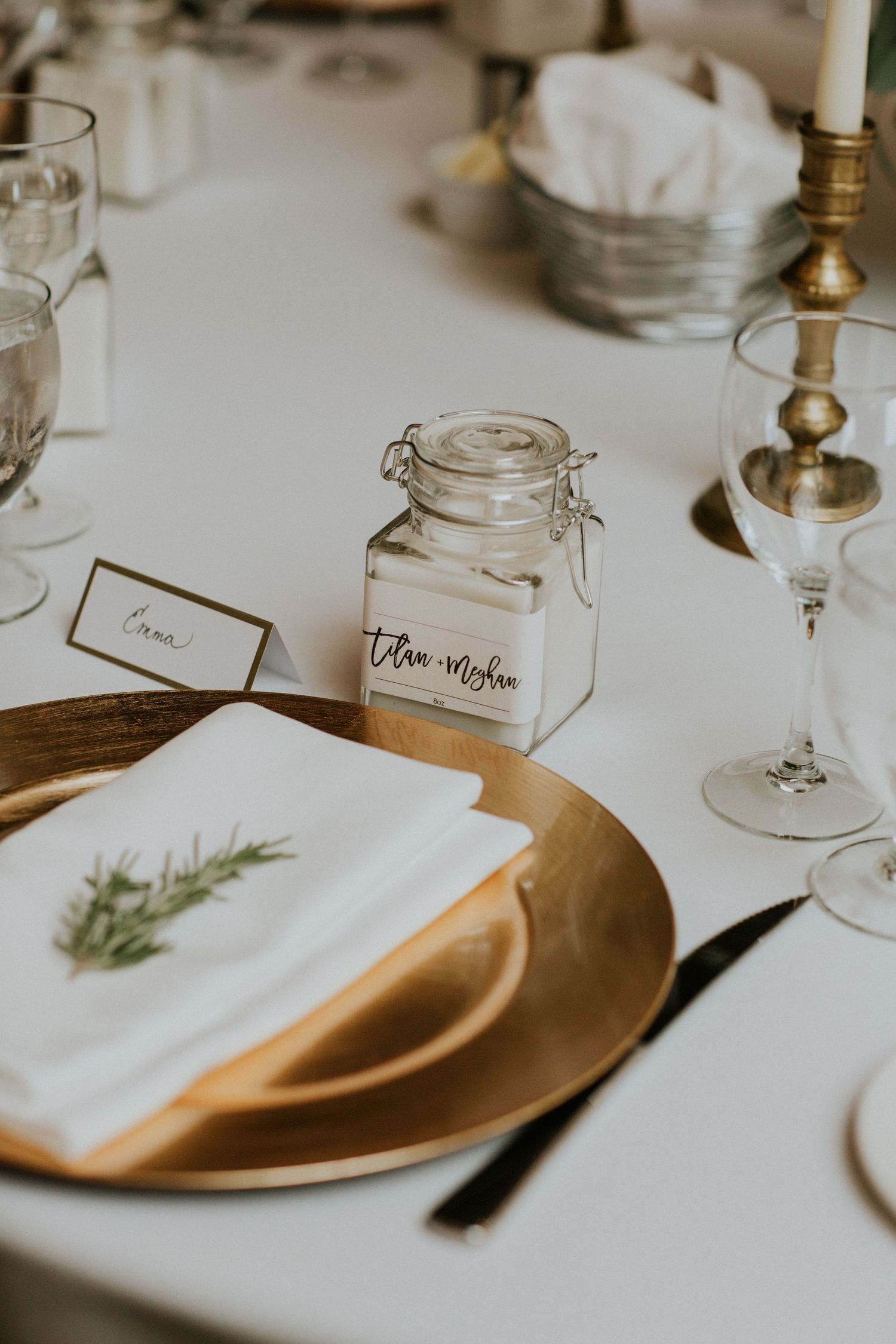 Elegant place setting with gold charger plate, white napkin, and glass jar centerpiece on white tablecloth.