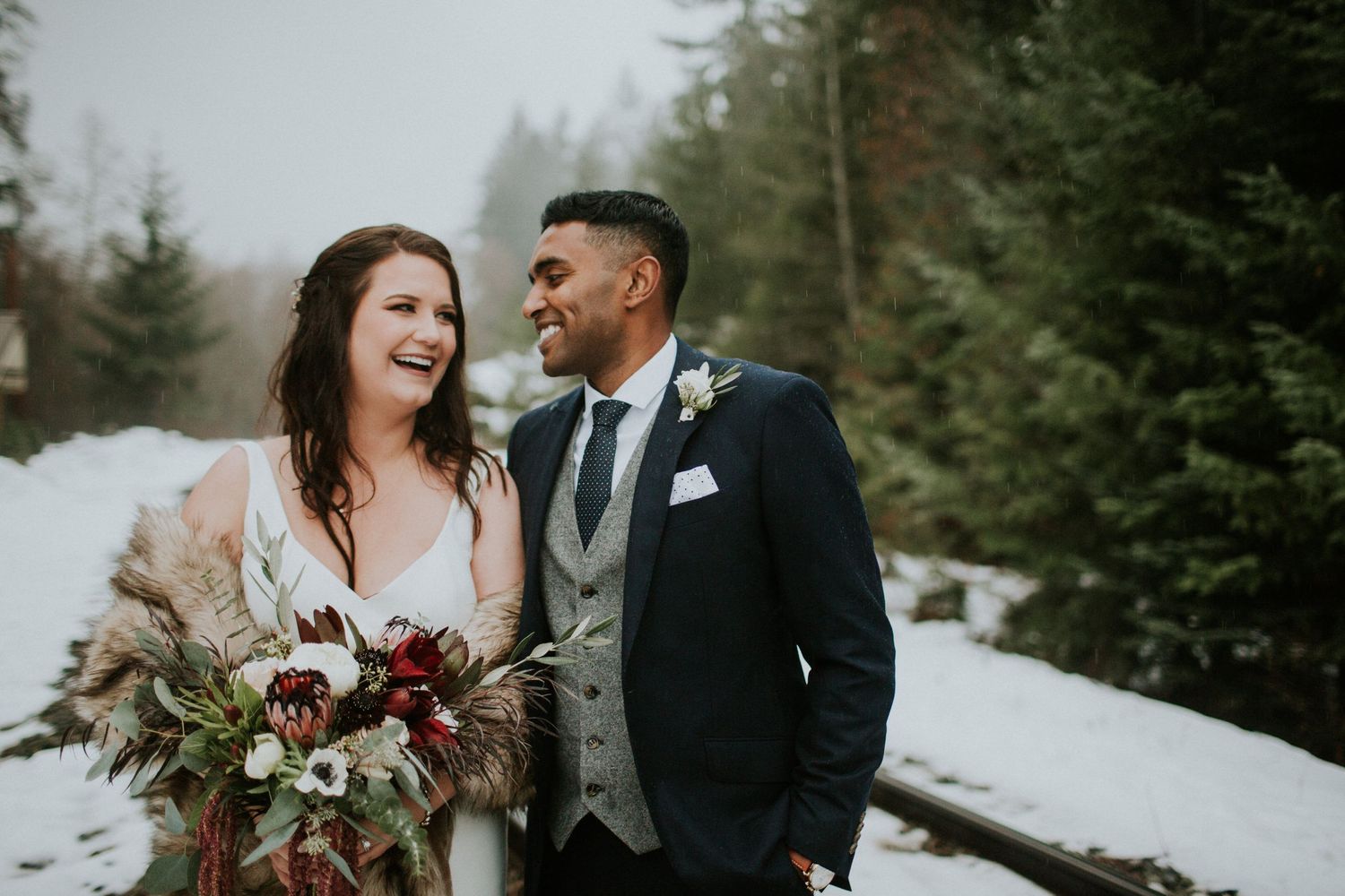 Winter wedding portrait along snowy railroad tracks surrounded by evergreen trees.