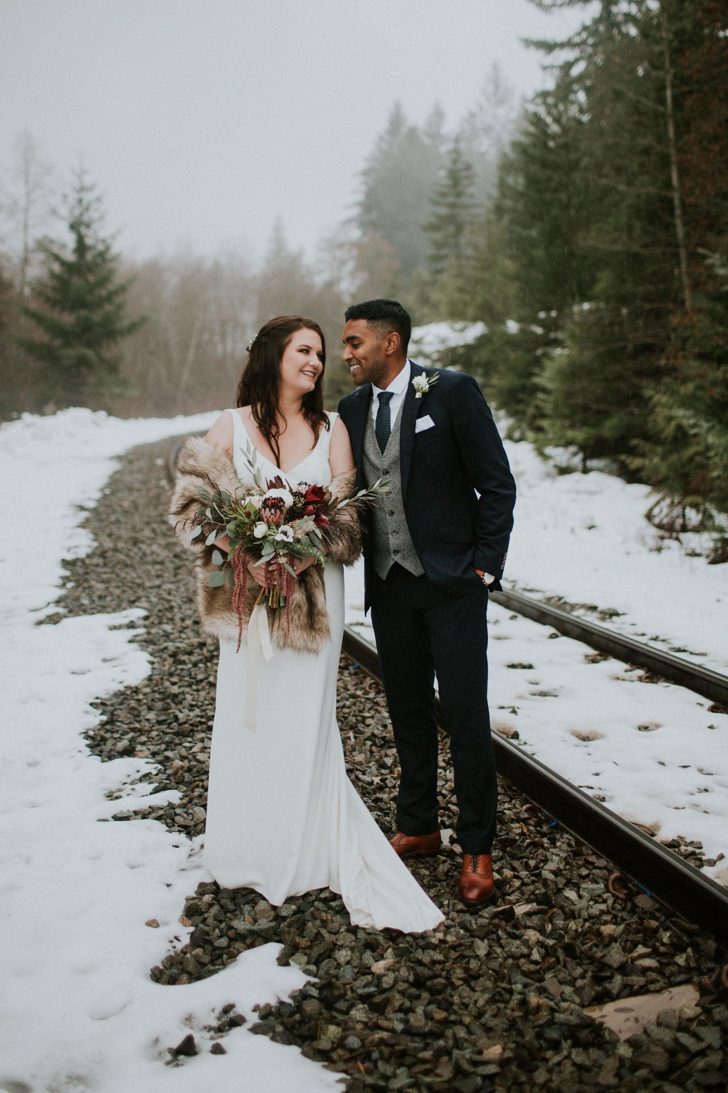 Intimate winter wedding moment on snow-covered train tracks with evergreen forest backdrop.
