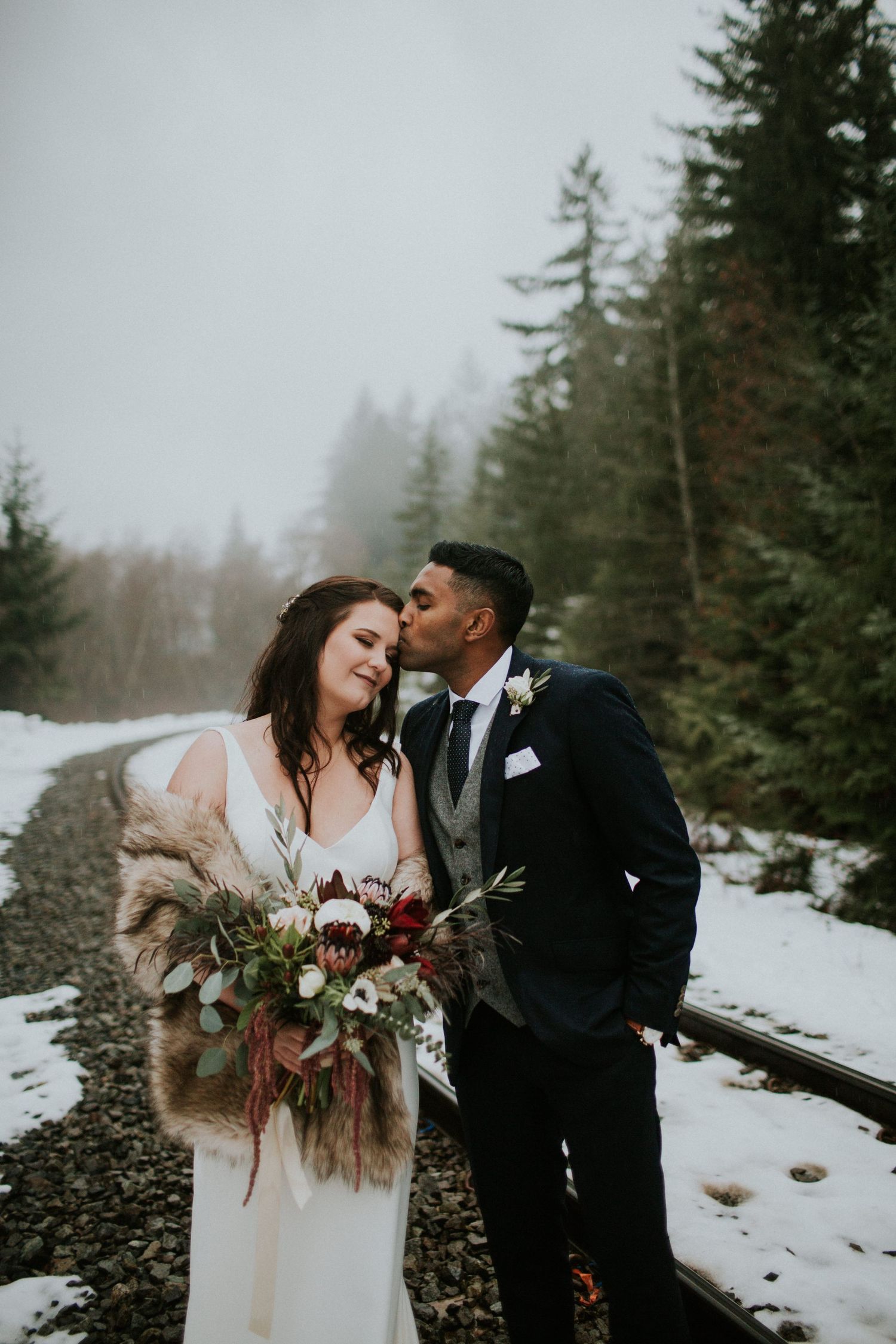 Tender winter wedding moment captured on snow-covered train tracks in foggy forest.