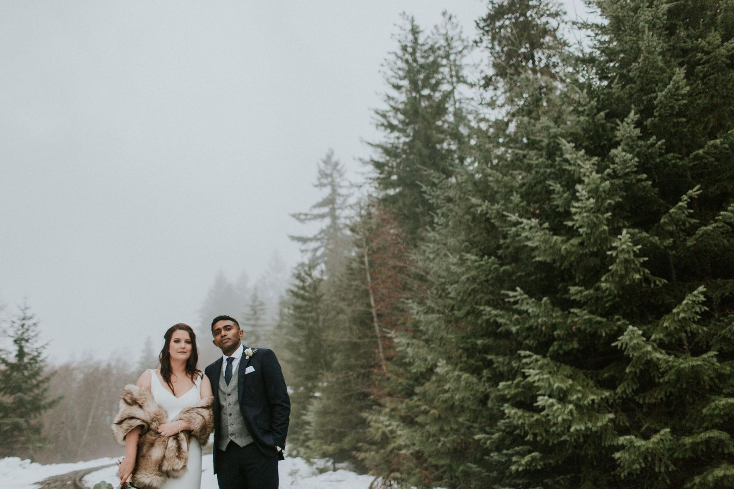 Winter wedding couple standing together on snowy railroad tracks surrounded by evergreens.