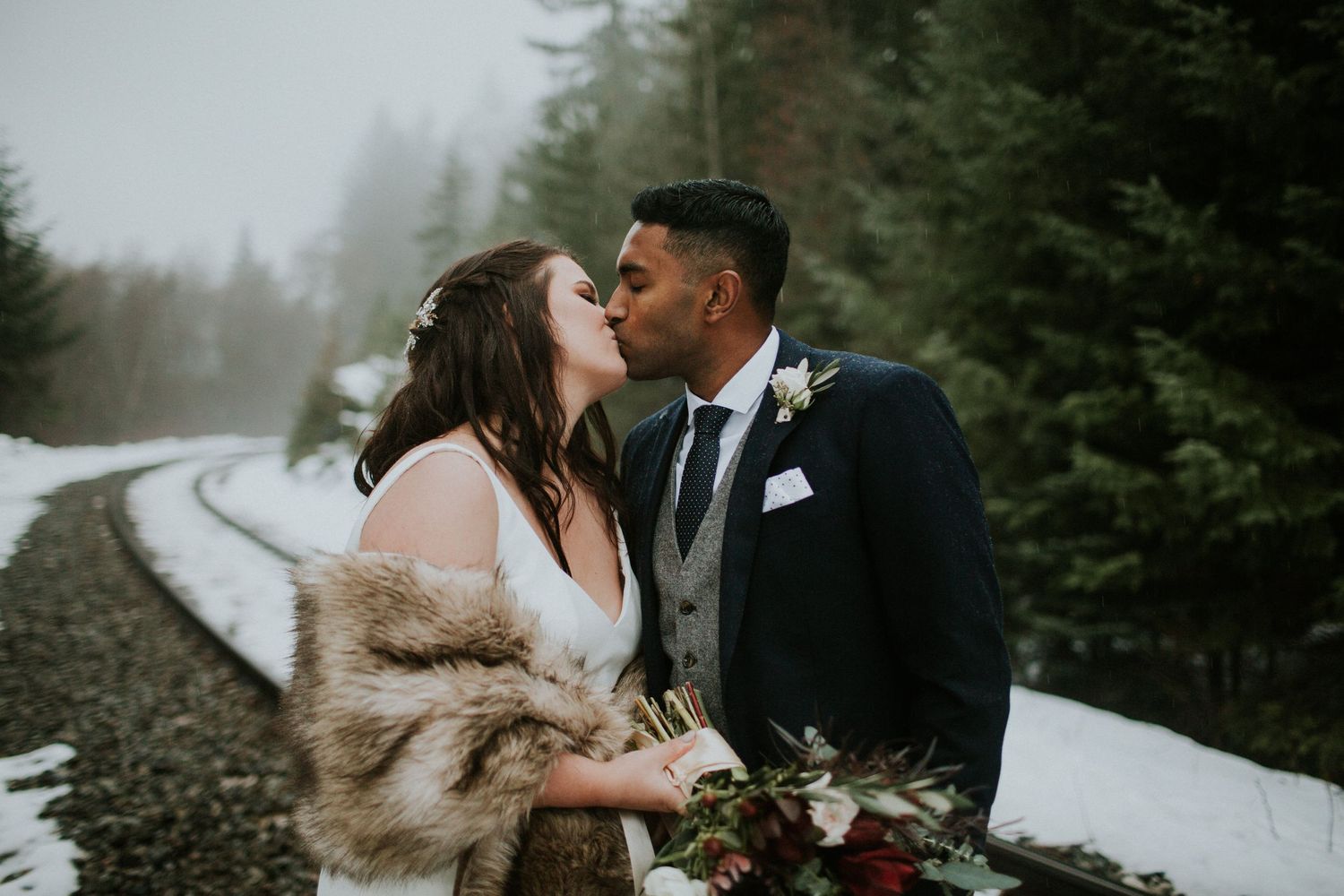 A romantic winter wedding photo on a snowy railroad track with evergreen trees in the background.