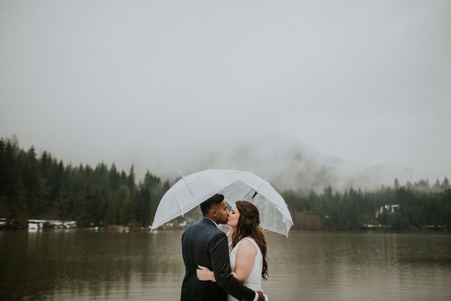 Wedding couple sharing an intimate moment under clear umbrellas by a misty lake.