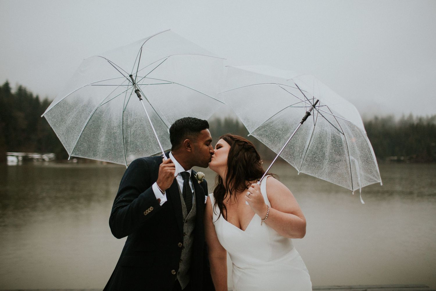 Newlyweds with clear umbrellas share a kiss in the rain with mountains in the background.