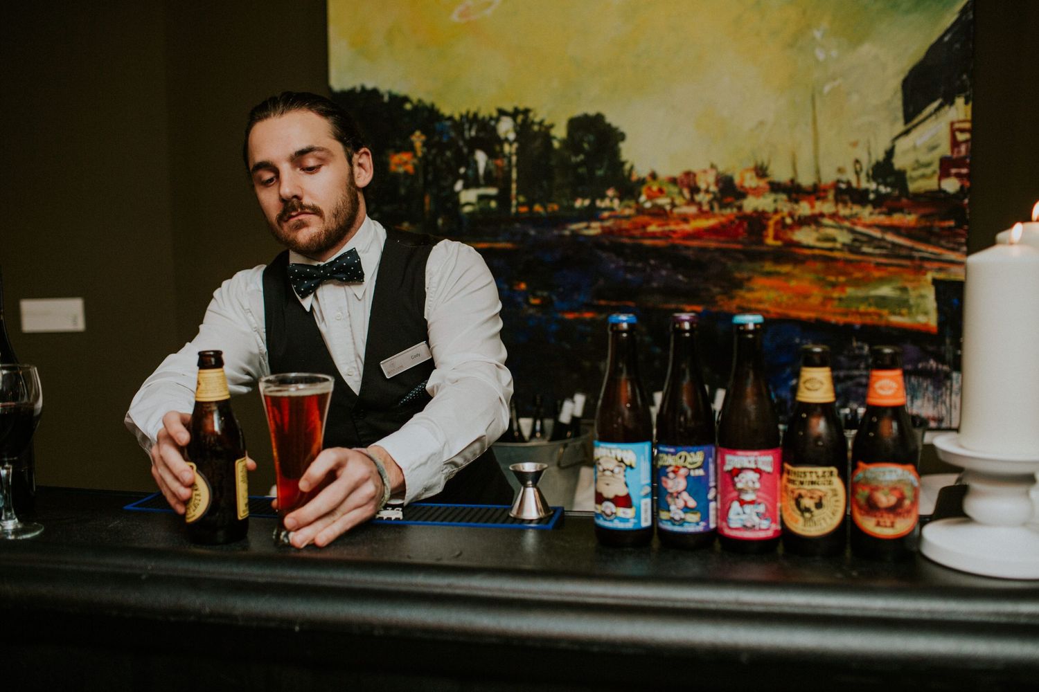 Bartender in a bow tie pouring craft beer from taps with colorful bottles displayed behind.