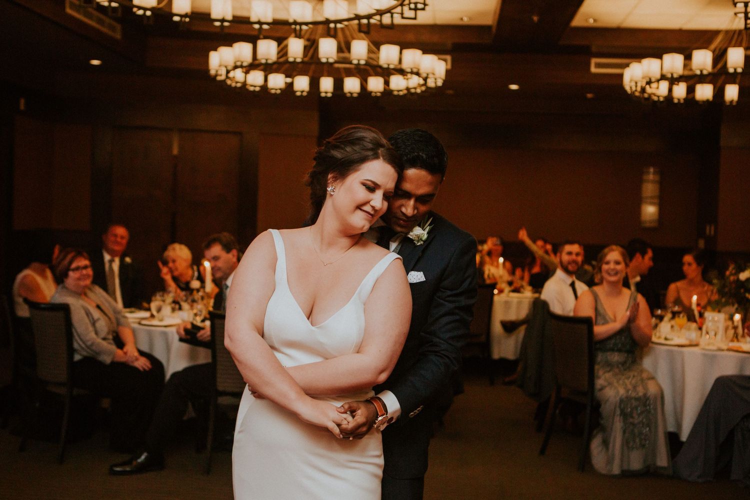 A couple shares their first dance at a wedding reception with guests seated at tables in the background.