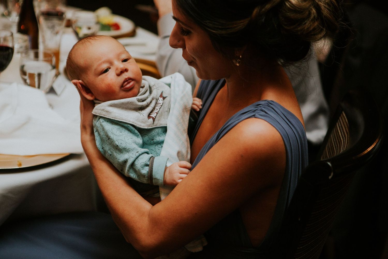 A baby in light blue clothing smiles while being held during a wedding reception.
