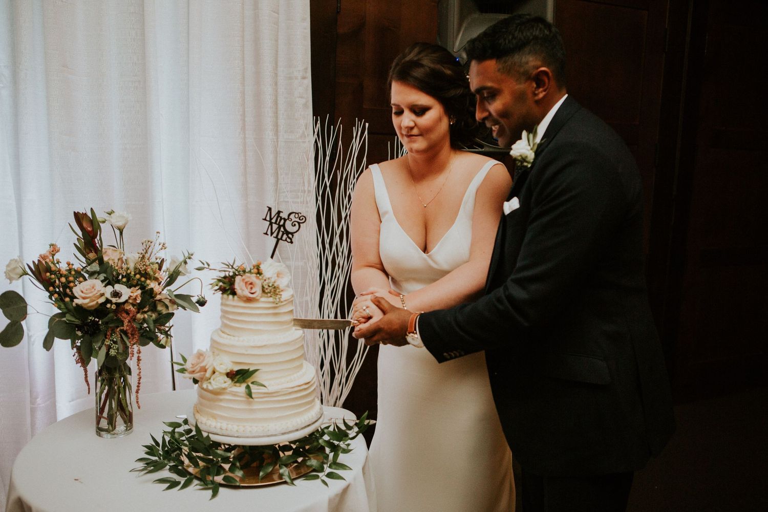 Newlyweds cut their elegant white wedding cake together beside floral arrangements.