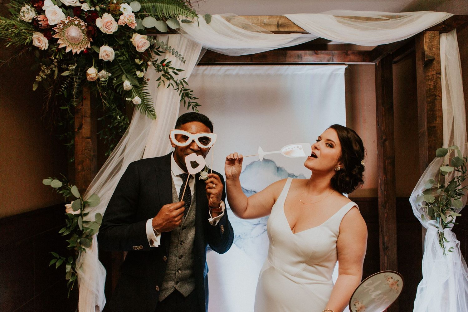 A playful wedding moment with drinks and masks under a floral arch decorated with white fabric and roses.