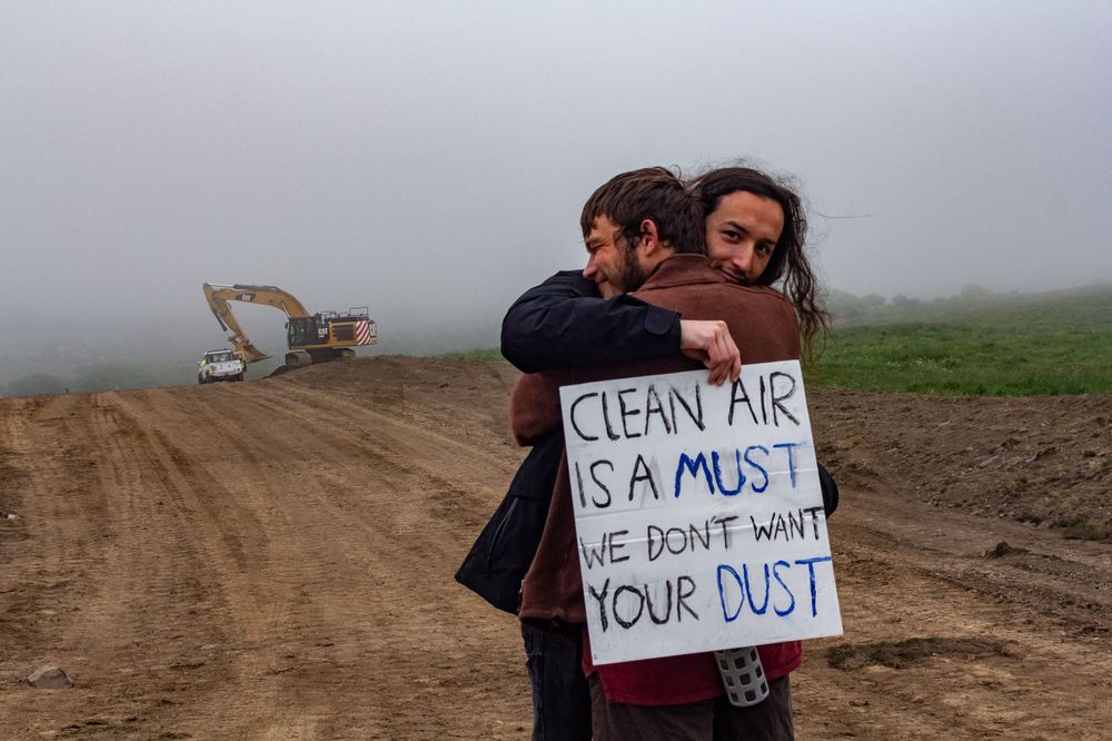 Two climate protectors hugging in front of a digger with a placard: "Clean air is a must, we don't want your dust."
