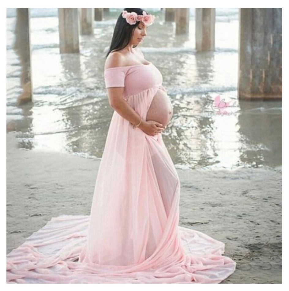 Pregnant woman in a pink off-shoulder chiffon maternity gown posing on a beach at sunset.