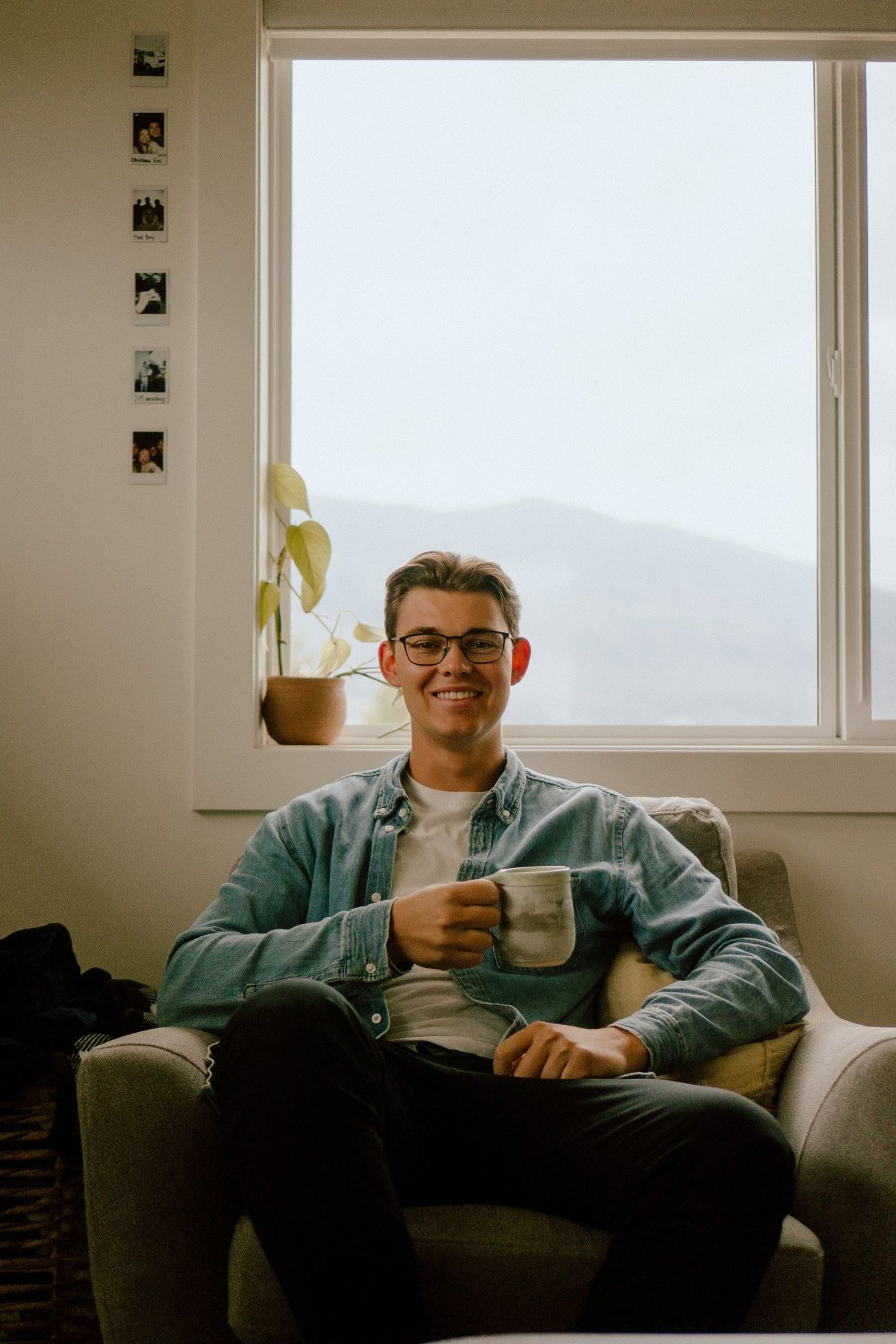 Person sitting in grey armchair by window holding coffee cup with photo gallery wall.