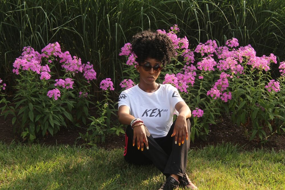 Person in white REX t-shirt crouches in front of vibrant pink flowering bushes in a garden setting.
