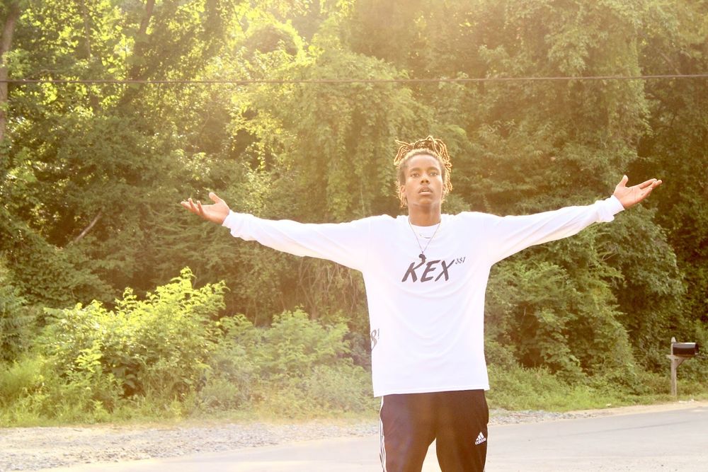 Person in white REX sweatshirt stands with arms outstretched against sunlit forest background.