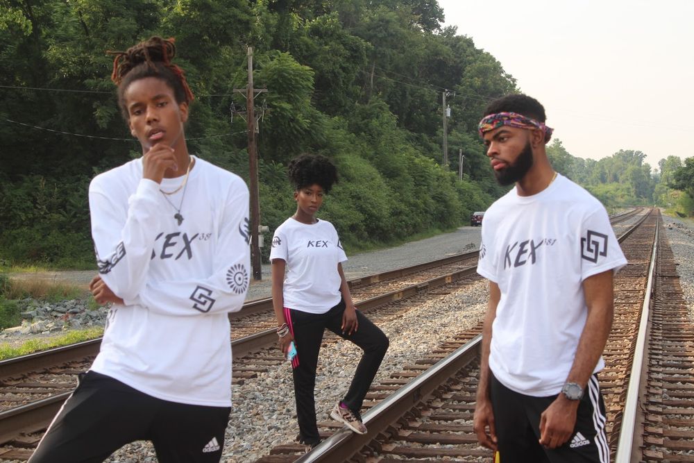 Three people in white REX shirts pose on railroad tracks with natural backdrop.