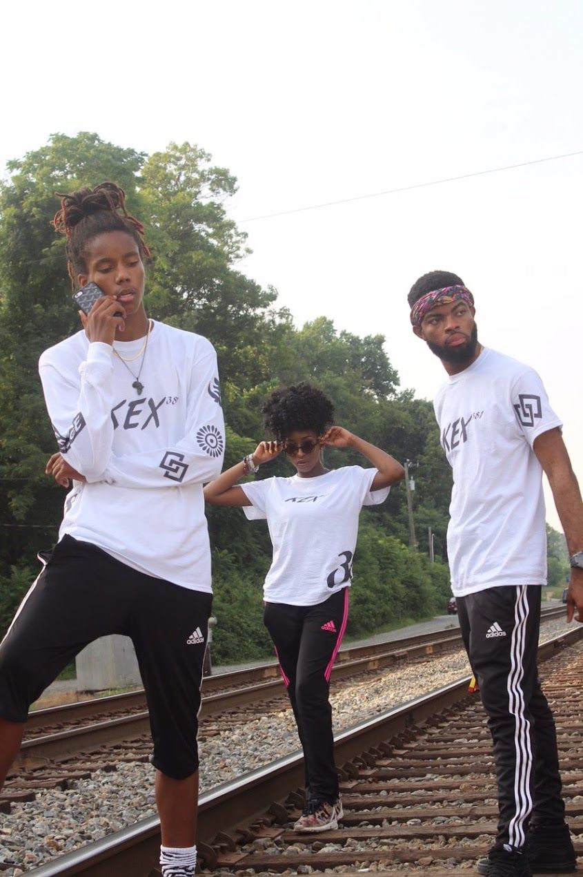 Three people in white REX shirts dance and pose on railroad tracks.