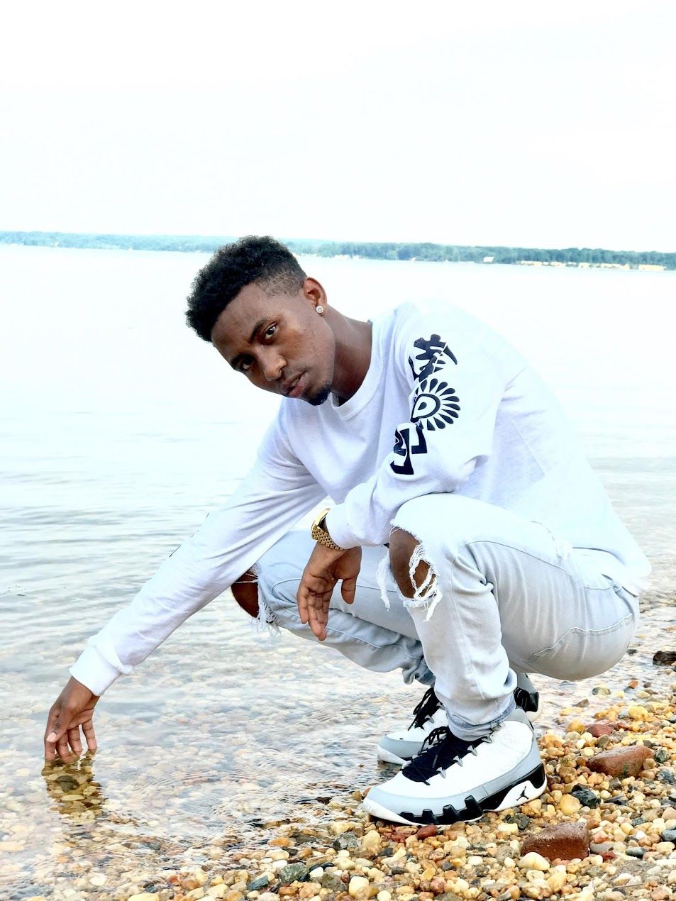Person in white REX sweatshirt crouches by waterfront in athletic pose.