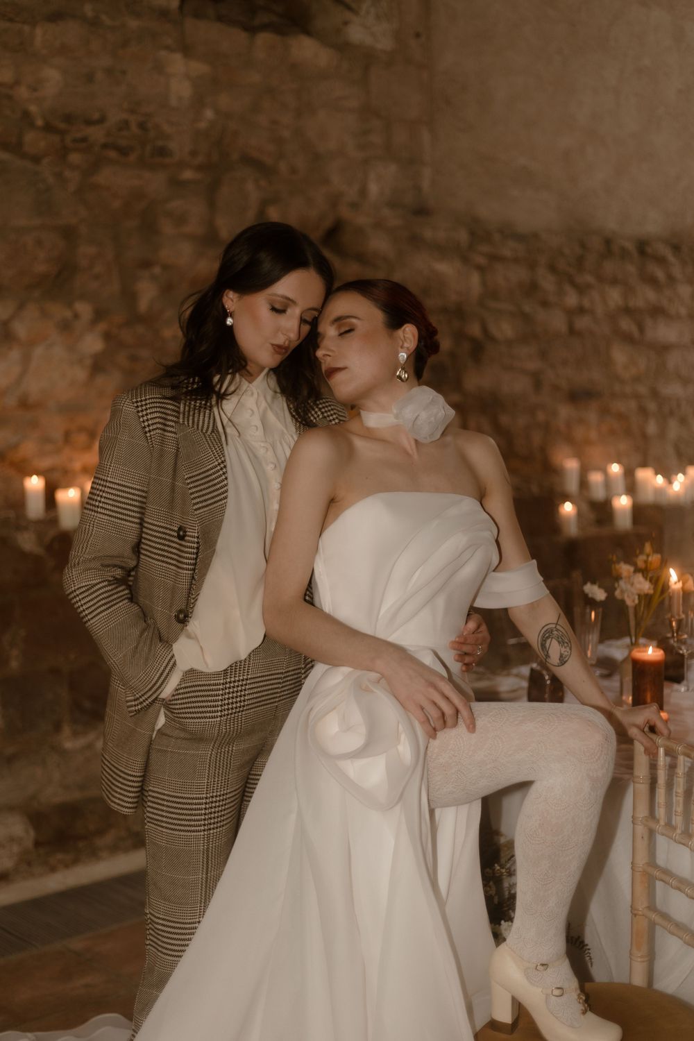 Editorial fashion shot of two models in bridal and suit attire with candlelit background.