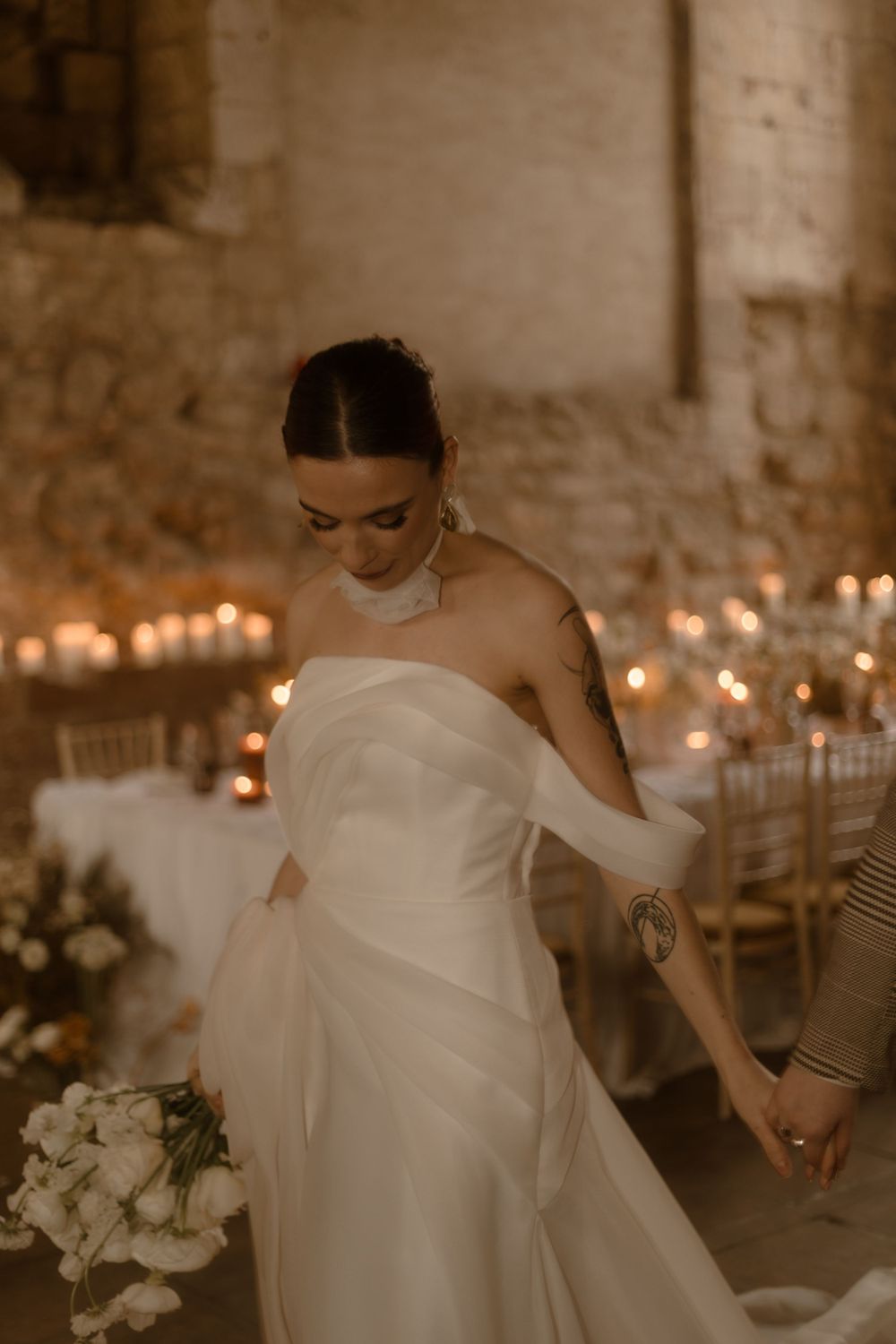 Modern bride in flowing white gown dancing amid candlelight in rustic stone venue.