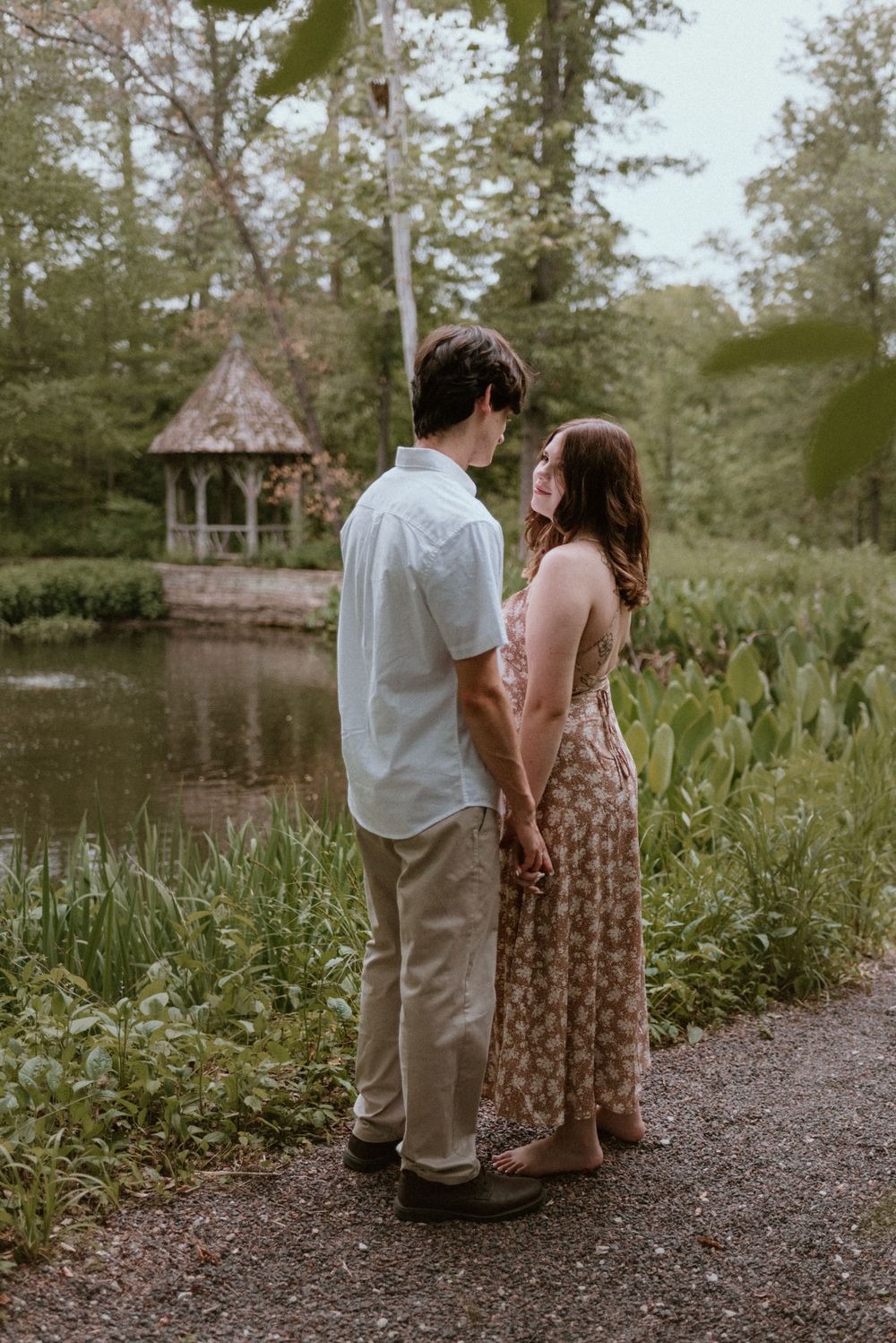 A couple strolls along a lakeside path near a gazebo at sunset.