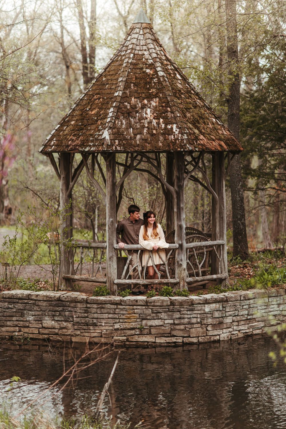 A rustic gazebo with a thatched roof sits on a stone wall overlooking a peaceful pond.
